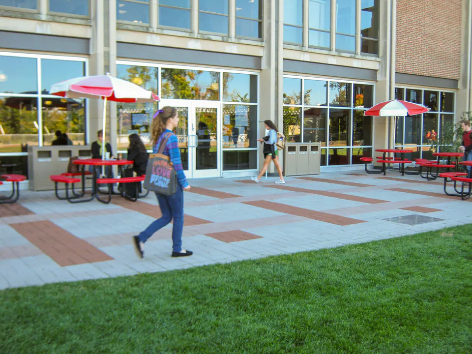 Campus building plaza with paver field, lawn edge, tables, umbrellas, and pedestrian traffic